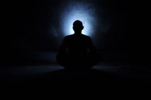 A man sitting in the dark with his hands folded on his chest.